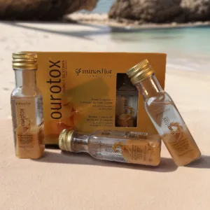 Muestra ampollas de botox capilar color oro, colocadas sobre la arena de una playa, al fondo se observa el mar.